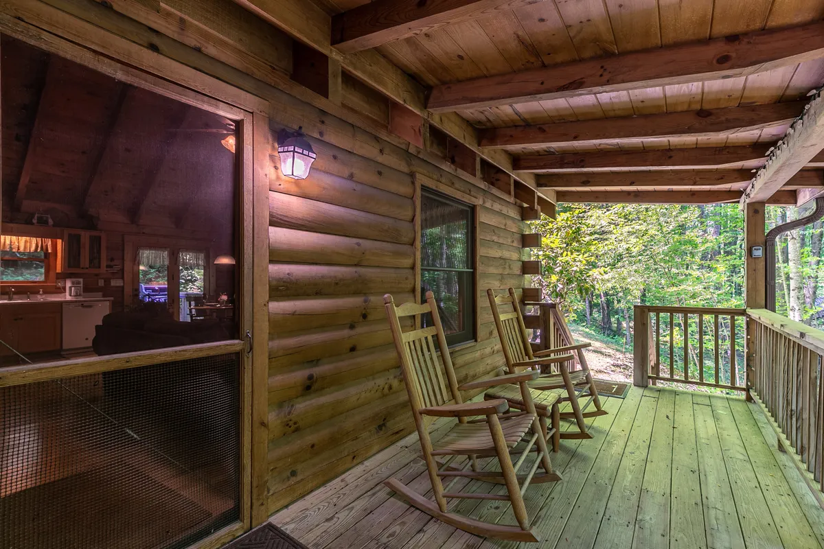 This image captures a portion of the covered deck, highlighting a pair of classic rocking chairs positioned to face the natural greenery. Wooden railings and the log cabin wall add rustic charm, while the leafy backdrop offers a sense of privacy and seclusion. The deck invites guests to relax, enjoy a morning coffee, or watch wildlife.