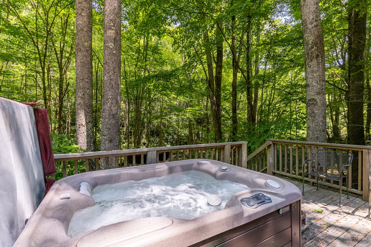 This image showcases the covered creekside deck with a hot tub nestled among lush trees. The hot tub sits prominently with steam gently rising, surrounded by comfortable seating and rustic wooden railings. The forested landscape creates a private, tranquil setting, and the gentle flow of the creek can be glimpsed beyond the deck. The overall ambiance is peaceful, inviting relaxation and connection with nature.