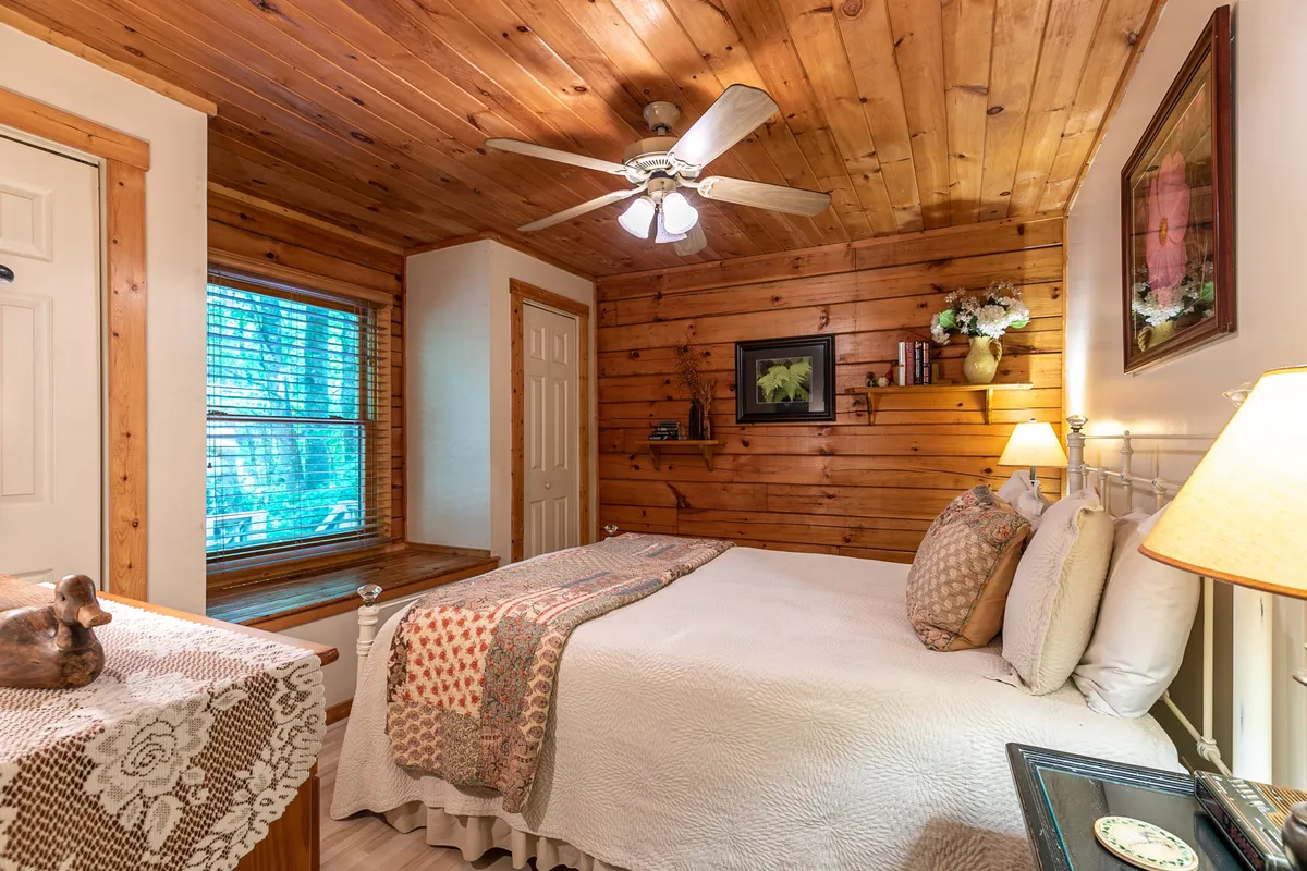 A tranquil bedroom scene with a wooden ceiling, a bed adorned with a decorative quilt, and natural light streaming through a window. A table lamp and decorative wall art add to the cozy ambiance. The window nook invites guests to wake up with nature, read, or unwind in peace.