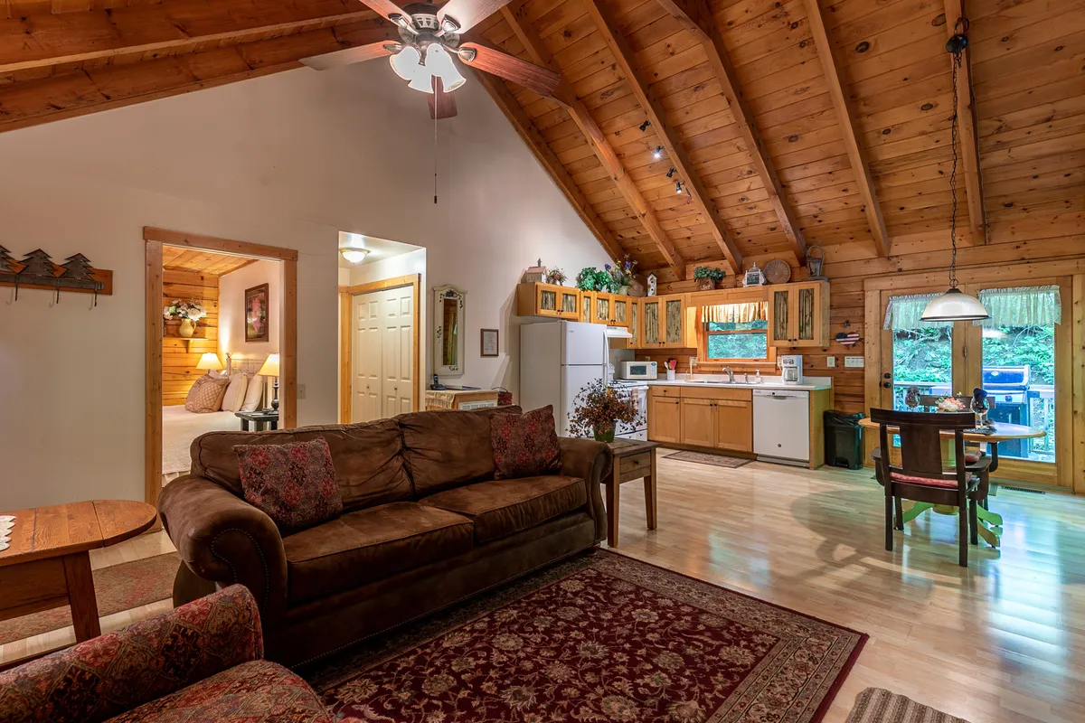 This image features a warm and welcoming interior that highlights a cozy sofa, a dining table set for two, and a view of the kitchen area. The space evokes togetherness and comfort, with an inviting atmosphere and natural light streaming in from large windows.