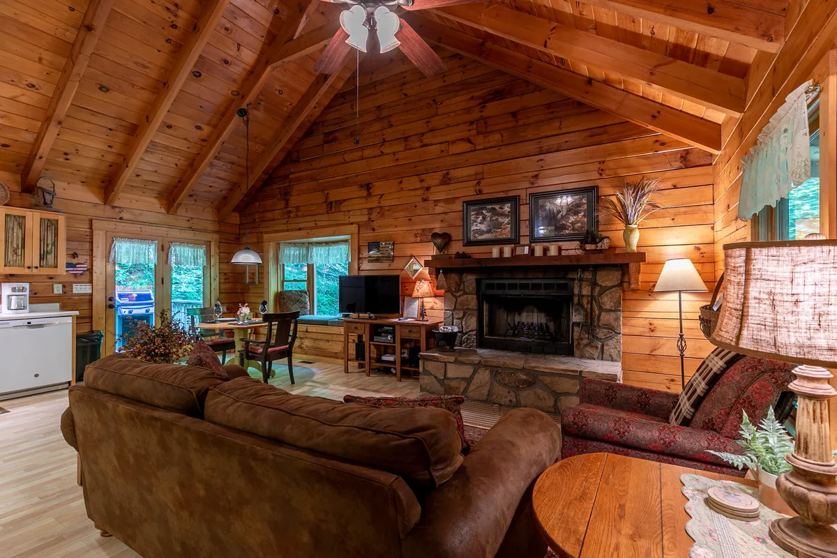 The open-plan interior reveals a stone fireplace, wooden beams, a comfortable sofa, and a dining area set for intimate meals. The ambiance is both warm and welcoming, perfect for relaxing by the fire, sharing conversations, or enjoying a cozy night in.