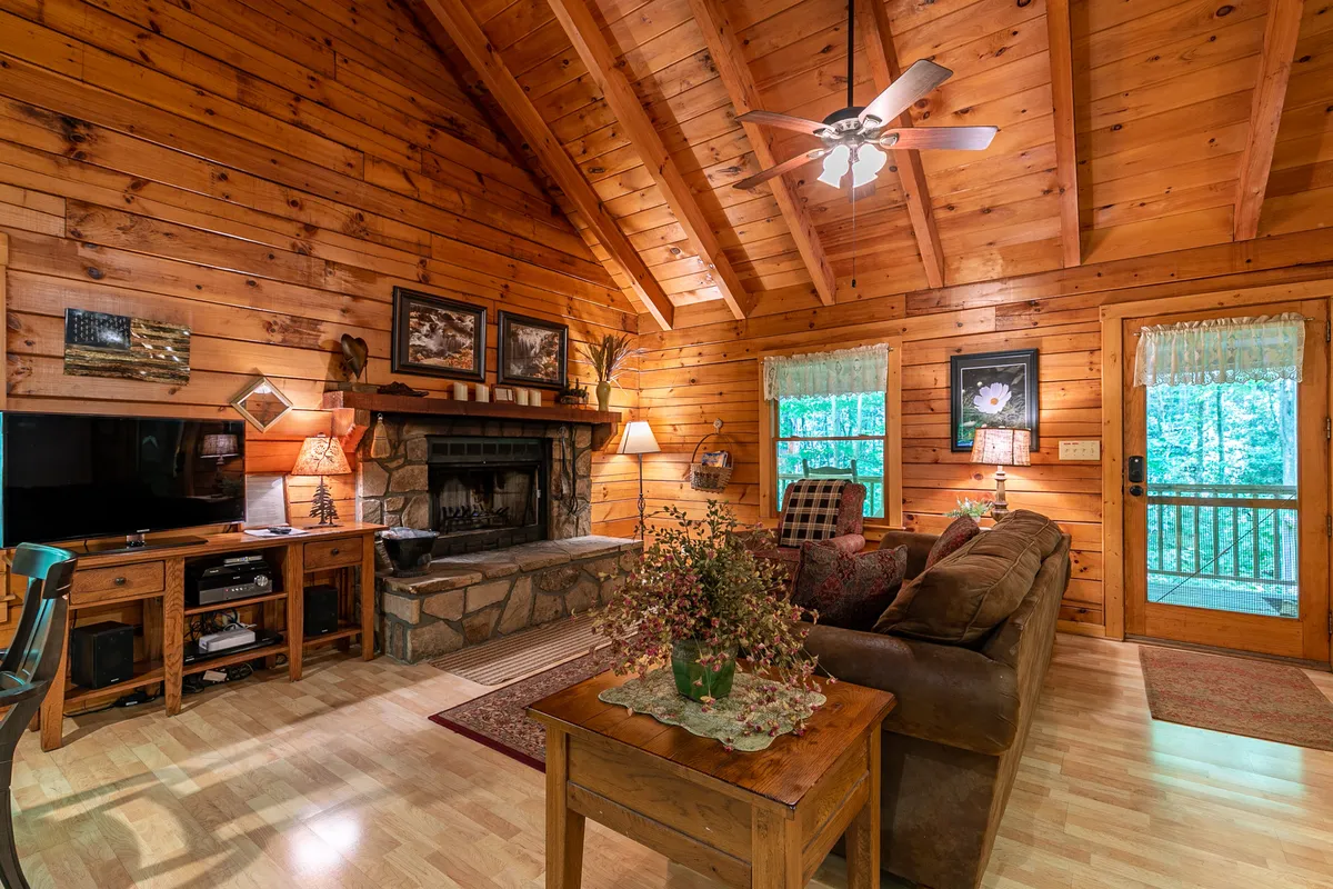 This inviting living area features a striking stone fireplace as its centerpiece, flanked by wooden beams overhead for a rustic touch. A comfortable sofa faces the fire, with a TV console and decorative artwork completing the cozy, open-plan space. Large windows provide scenic views and allow natural light to pour in, enhancing the warmth and atmosphere.
