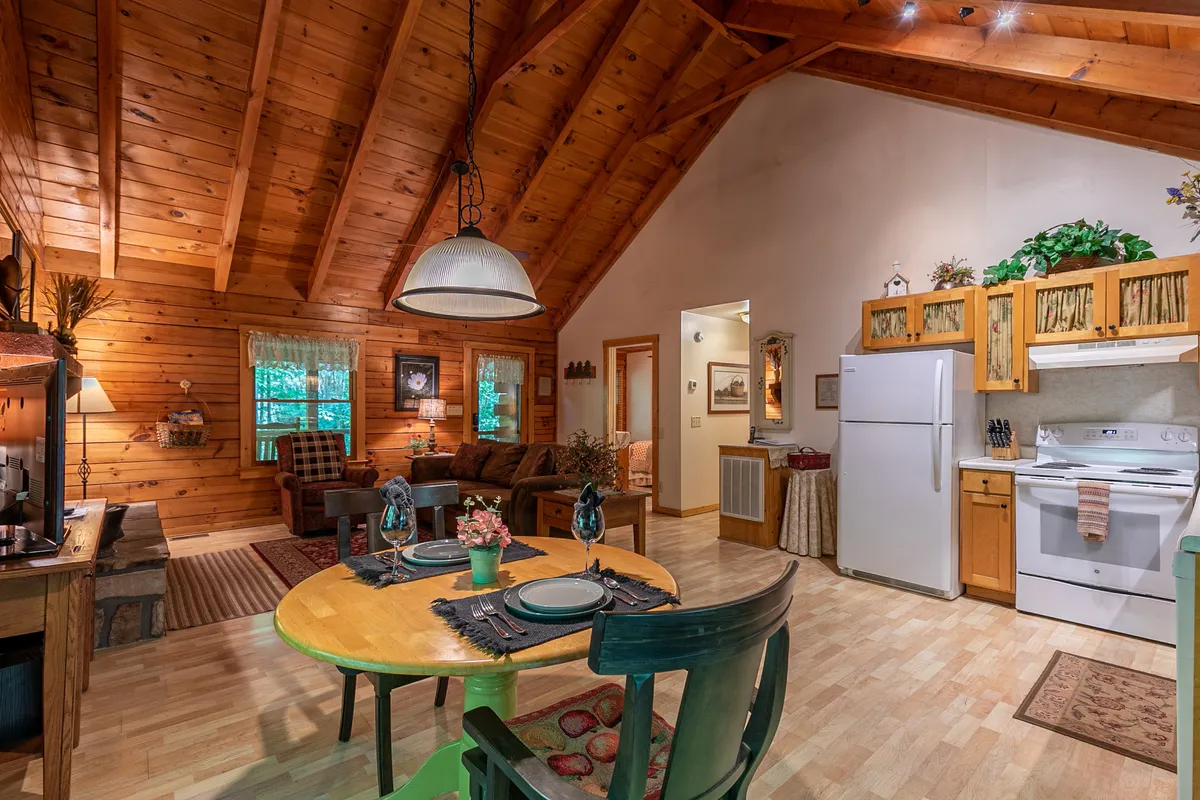This image presents the heart of the cabin: a living and dining area anchored by exposed wooden beams, a family-style dining table, a plush sofa, and an inviting kitchen. The space exudes warmth and togetherness, signaling opportunities for shared meals, relaxing by the fire, and engaging in cozy group activities.