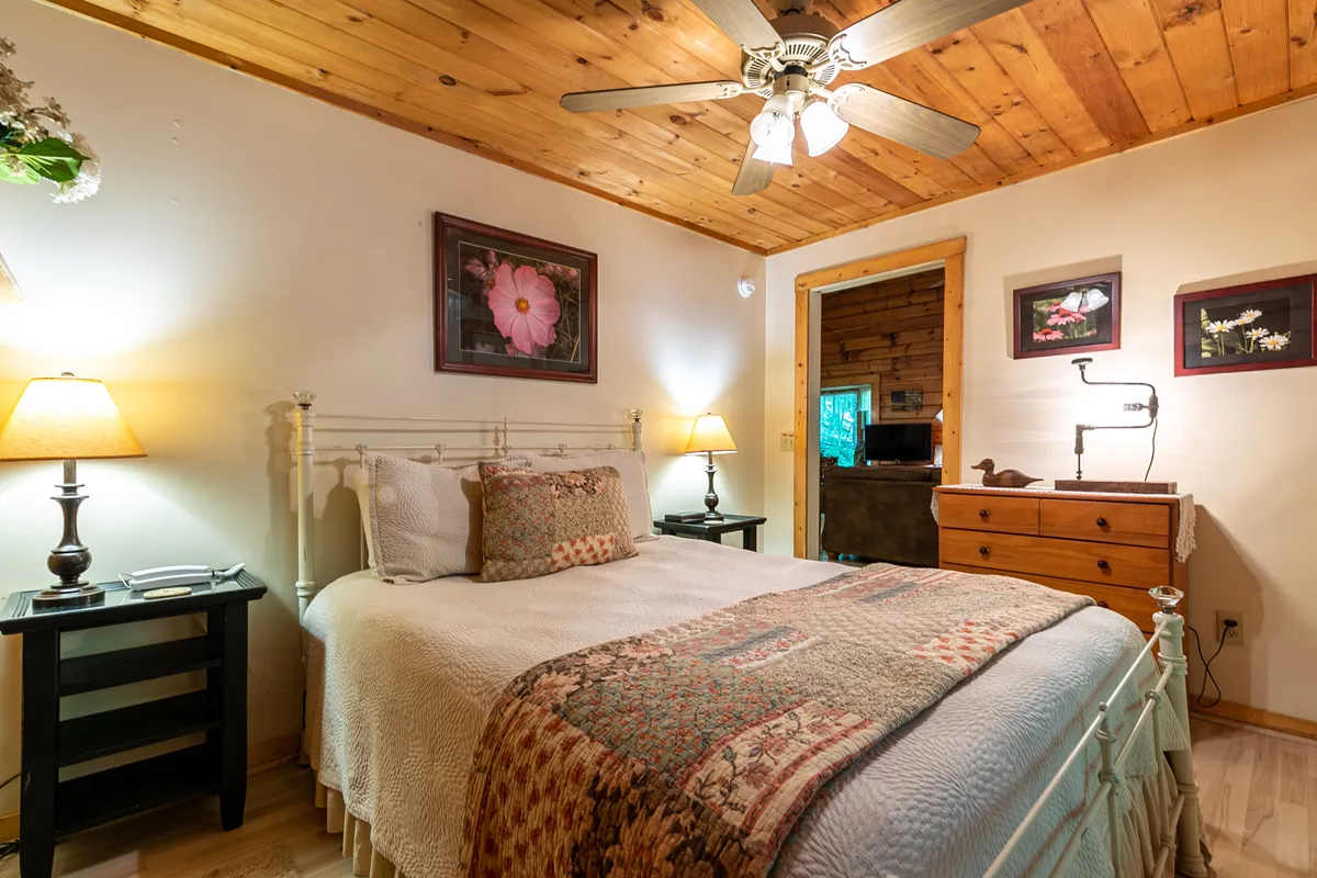 A serene bedroom showcases a plush Queen bed with crisp linens, flanked by nightstands and soft lighting. Artwork and a dresser add personal touches, while natural light streams in through windows, creating a peaceful haven ideal for rest and quiet moments.