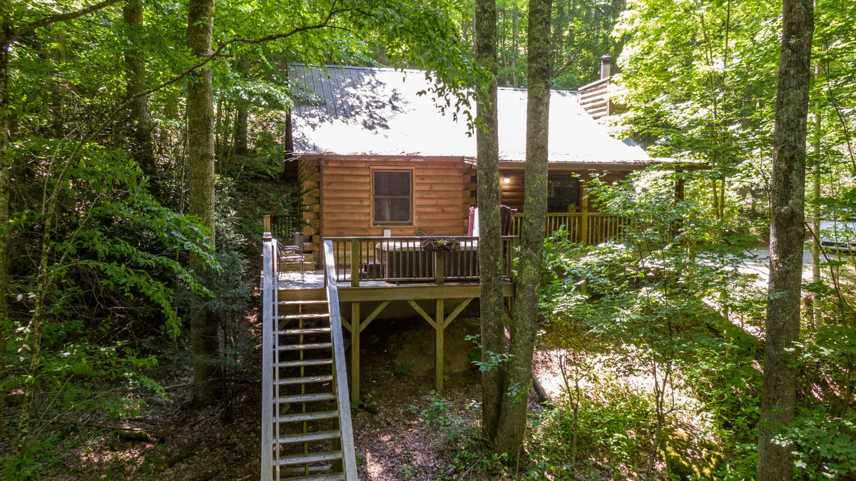 This image features the inviting log cabin set amidst lush greenery, with a prominent deck and wooden stairs leading to the entrance. The expansive outdoor area is surrounded by trees, offering a sense of privacy and immersion in nature. The deck space is ideal for relaxing, enjoying morning coffee, or sharing tranquil moments together.